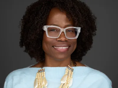 Professional woman with natural curly hair and decorative glasses in light blue top with statement necklace