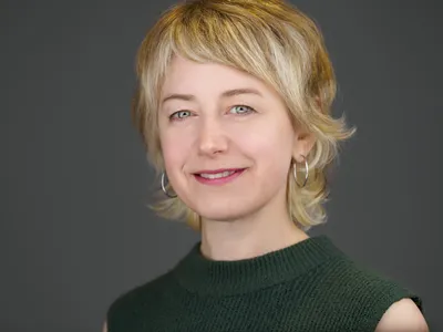 Professional woman with short blonde hair and fringe in forest green sleeveless top, subtle smile and hoop earrings
