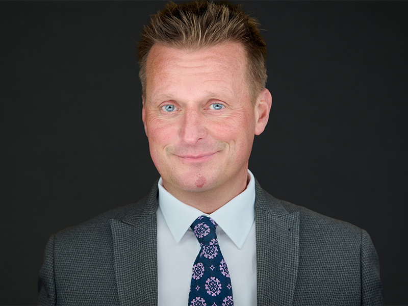 Professional corporate headshot of a business leader in a dark suit and tie against a dark background