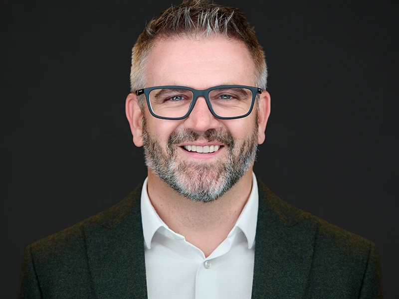 Corporate headshot of a professional man with glasses and a beard wearing a dark jacket and white shirt against a dark background