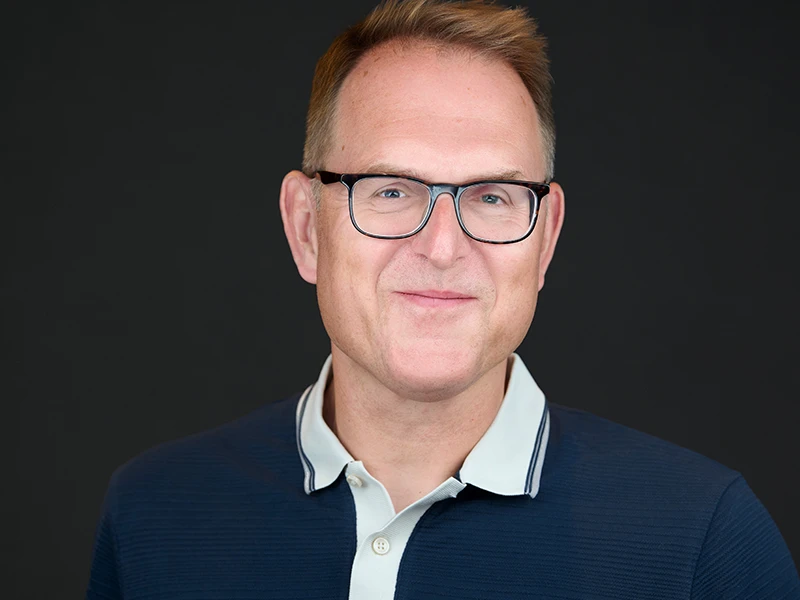 Professional corporate headshot of a man wearing glasses and a navy blue polo shirt against a dark background
