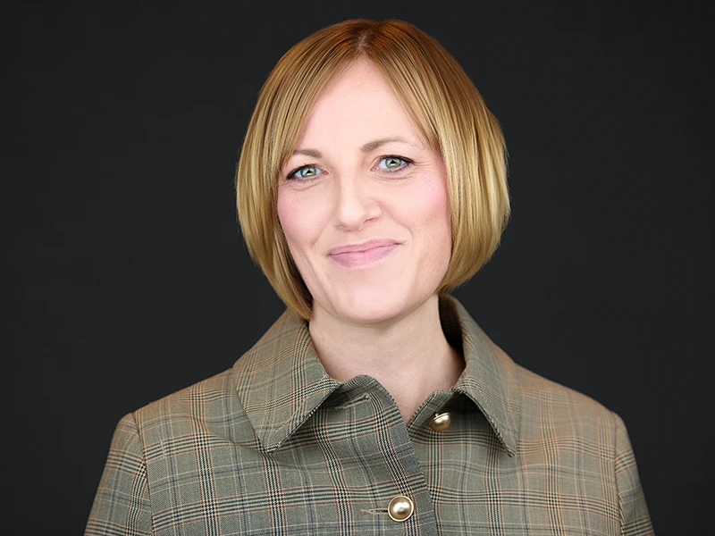 Corporate headshot of a professional woman with short blonde hair wearing a green jacket against a dark background