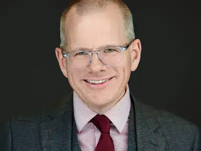 Corporate headshot of senior executive with glasses wearing dark suit and burgundy tie, showcasing professional leadership presence for corporate team photography