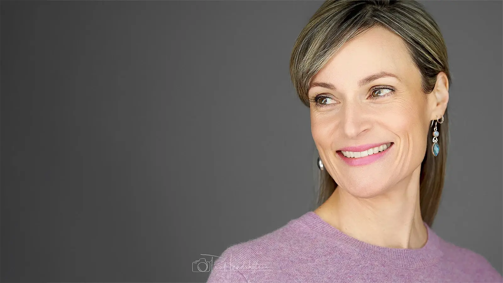 Exeter Headshots: Elegant Personal Branding Professional headshot of a woman wearing a mauve sweater and blue drop earrings, showing a natural, engaging smile against a dark background.