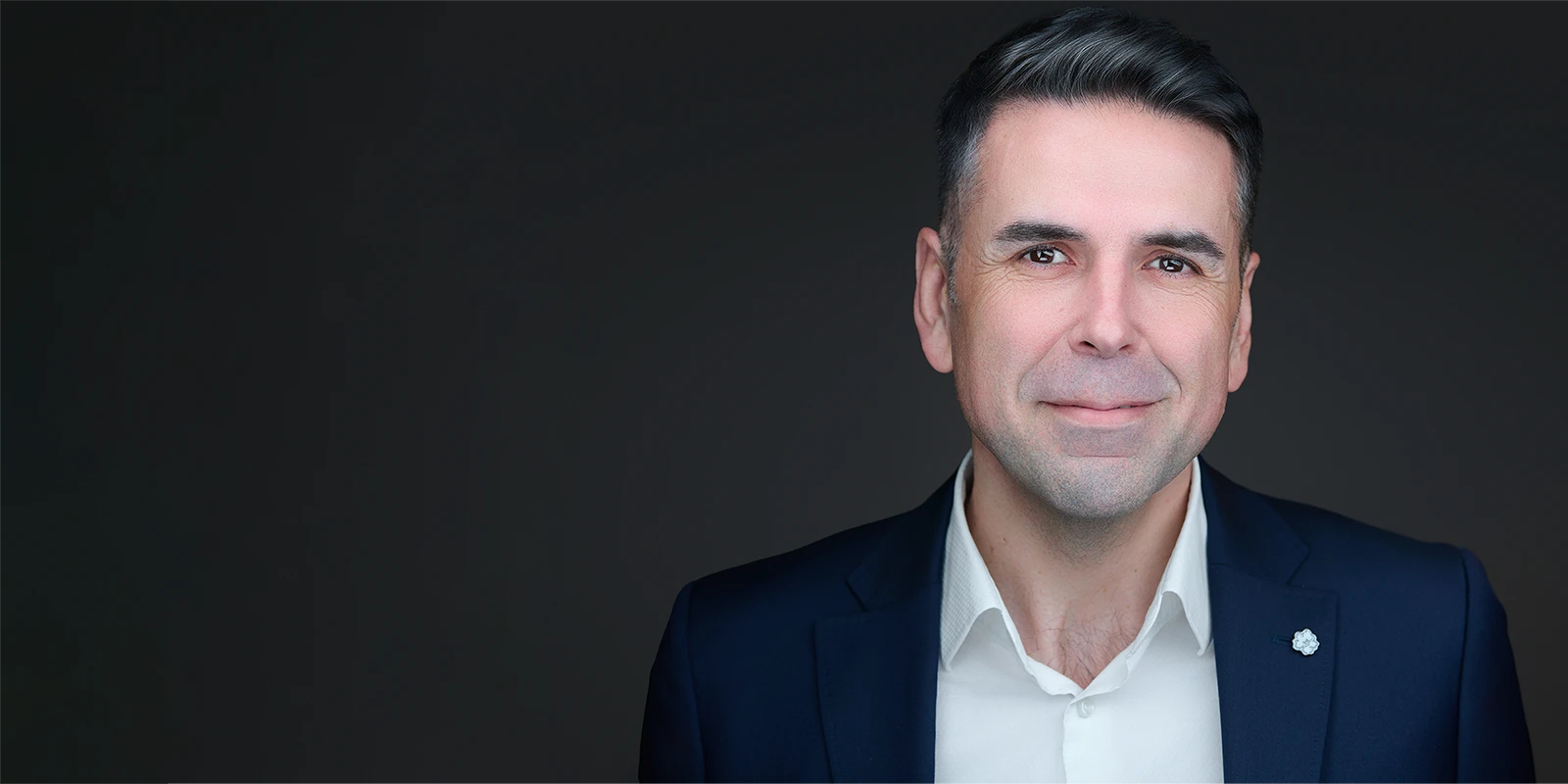 Professional corporate headshot of a man in a navy suit and white shirt on a dark background, with space for headline text.