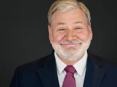 Smiling senior man with white beard in navy suit and burgundy tie, professional headshot on dark background.