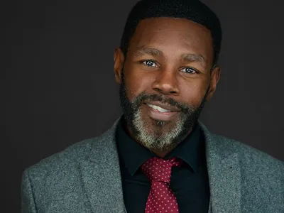 Professional headshot of a confident man with short hair and beard, wearing a grey blazer, black shirt, and red patterned tie, smiling against a dark background.