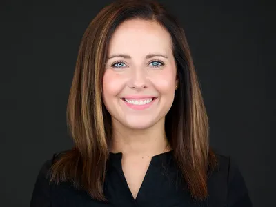 Smiling woman with straight brown hair in black top, professional headshot on dark background.