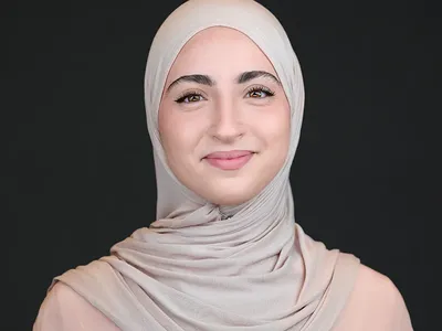 Smiling young woman wearing a light beige hijab, professional headshot on dark background.