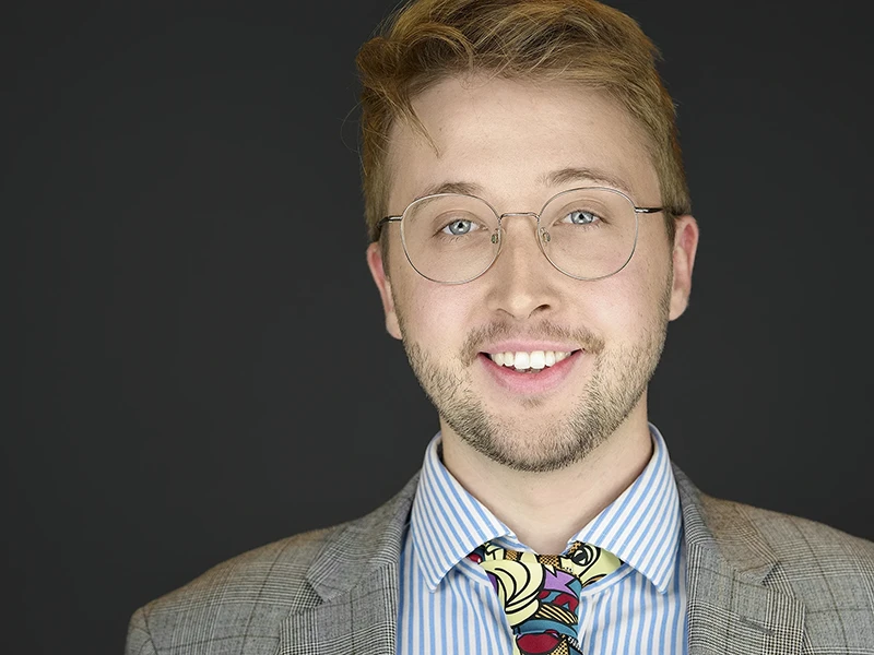 Professional headshot of a young man wearing round glasses, a grey check blazer, striped shirt and colourful patterned tie, showing a warm smile against a dark background.