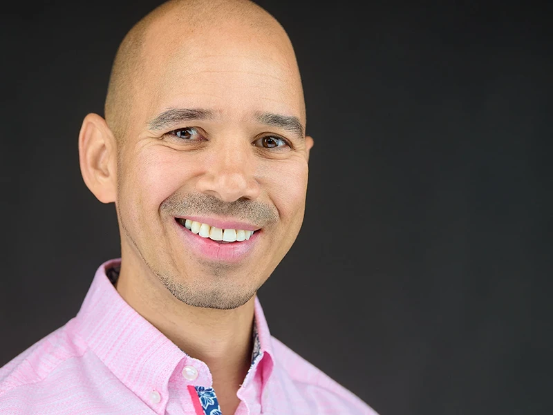 Professional headshot of a man wearing a pink button-down shirt with patterned collar detail, showing a bright genuine smile against a dark background. Pro Headshots logo visible.