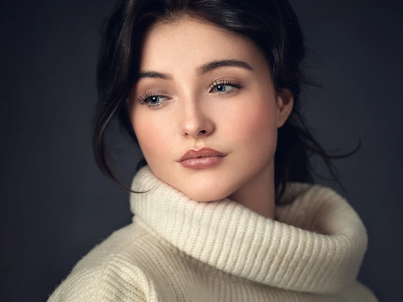 Professional headshot of a woman with dark hair wearing a cream turtleneck sweater, captured in a soft, elegant style against a dark background with gentle lighting highlighting facial features.