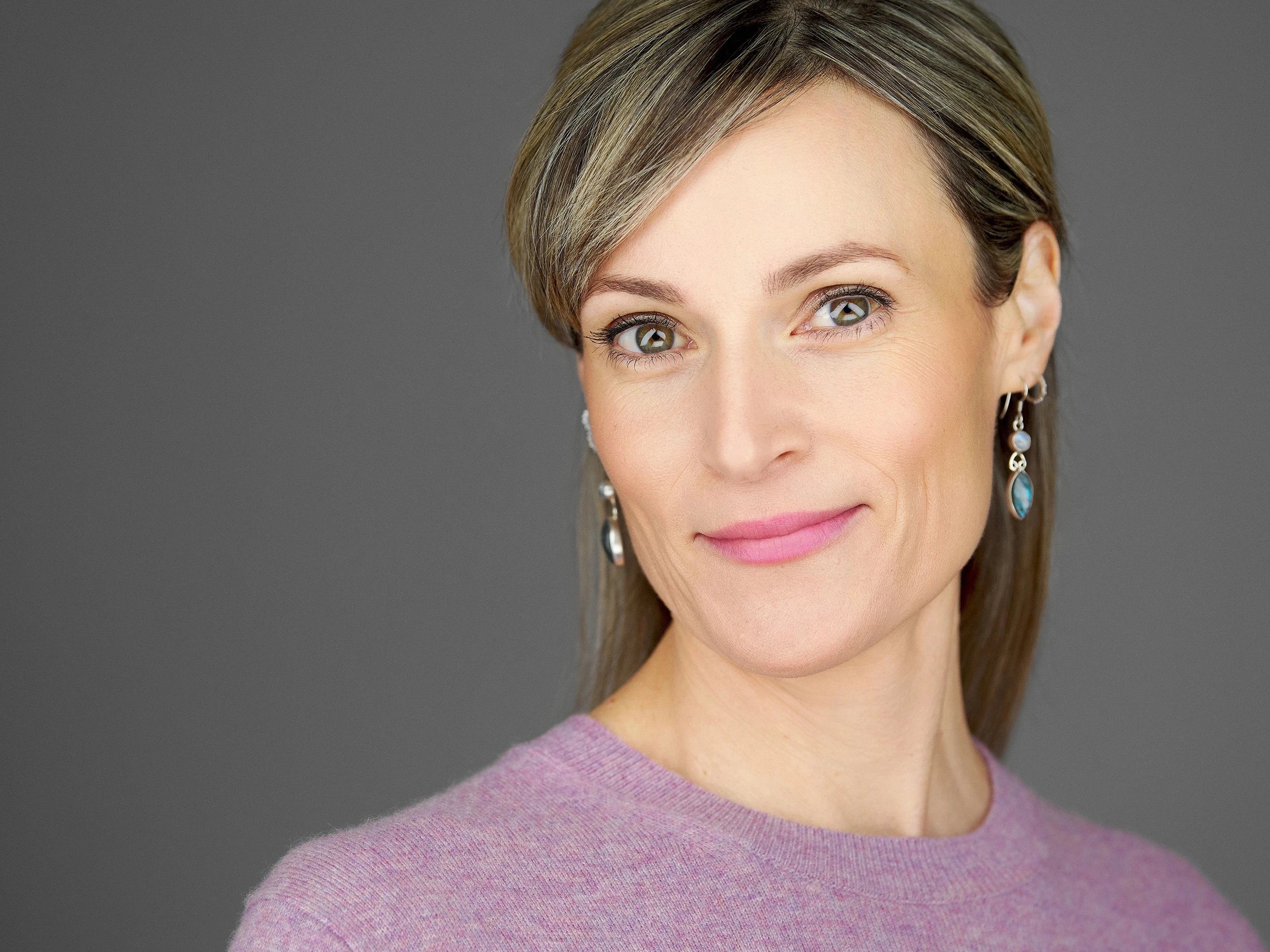 A woman wearing a pink jumper and turquoise drop earrings, with sleek brown hair styled with a side fringe, offering a subtle smile against a grey background. The portrait demonstrates refined headshot photography in Bristol with soft, flattering lighting.