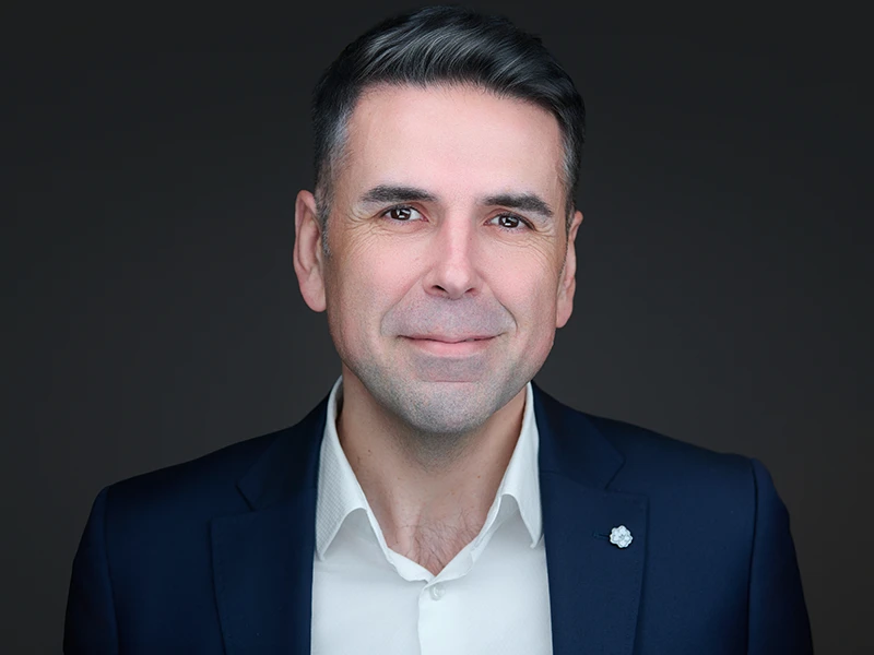 Smiling man with a beard in a light blue shirt, photographed on a dark grey background.