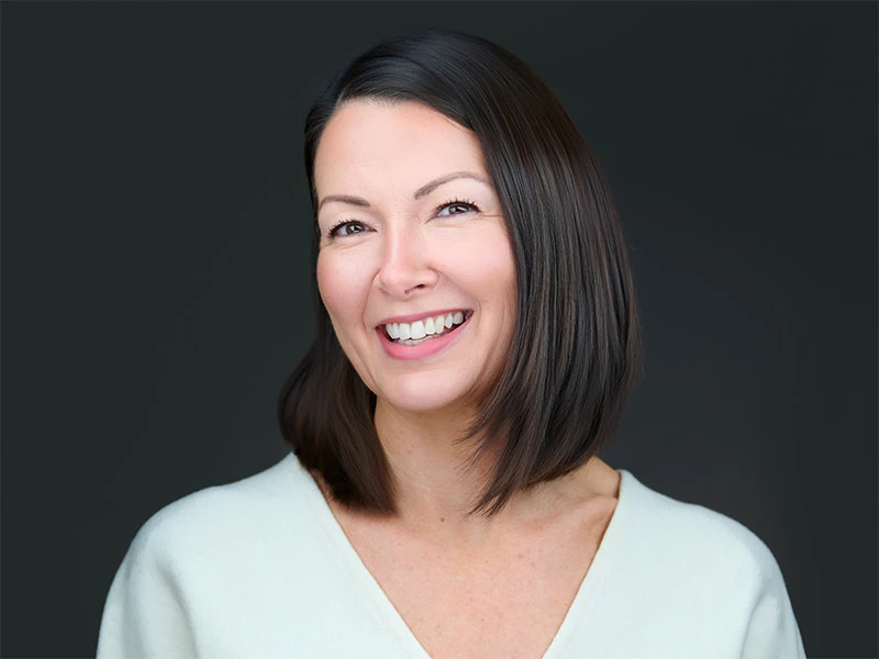 Smiling woman with a dark bob haircut in a white top, photographed on a dark grey background.