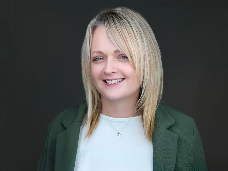 Smiling blonde woman in a green blazer and white top, photographed on a dark grey background.