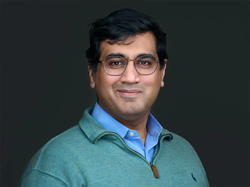 Man wearing glasses in a green quarter-zip jumper and blue shirt, photographed on a dark grey background.