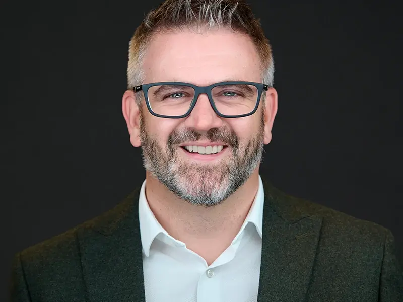 Smiling man with salt-and-pepper beard, dark-rimmed glasses and white shirt in green blazer, professional headshot on dark background.