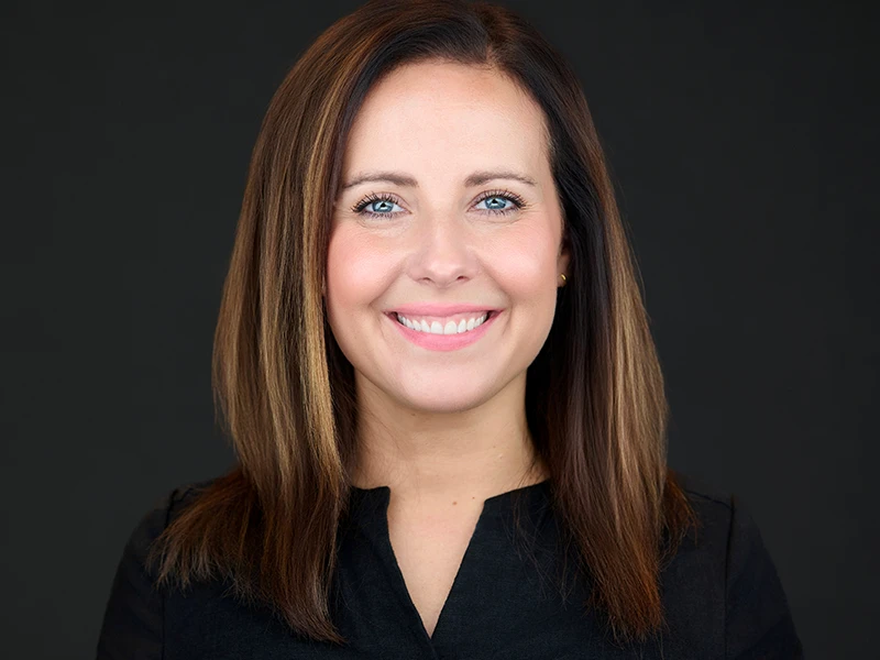 Smiling woman with straight brown hair in black top, professional headshot on dark background.