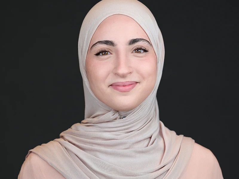Smiling young woman wearing a light beige hijab, professional headshot on dark background.