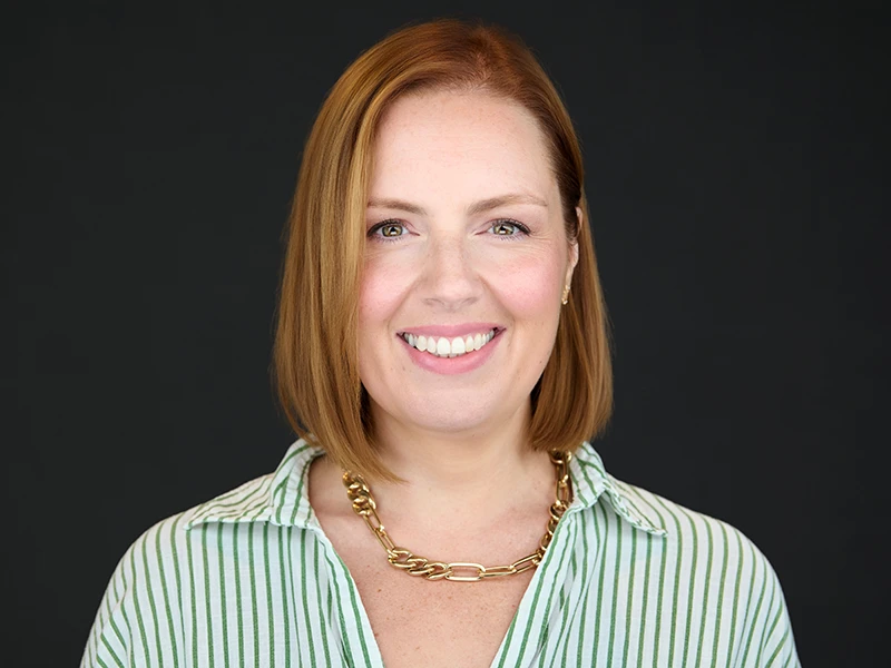 Smiling professional woman with shoulder-length auburn hair wearing a green-striped shirt and chunky gold necklace, photographed on a dark background.