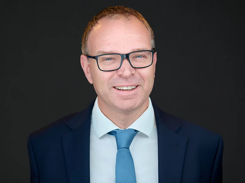 Smiling professional man in glasses wearing a navy suit and light blue tie, photographed on a dark background.