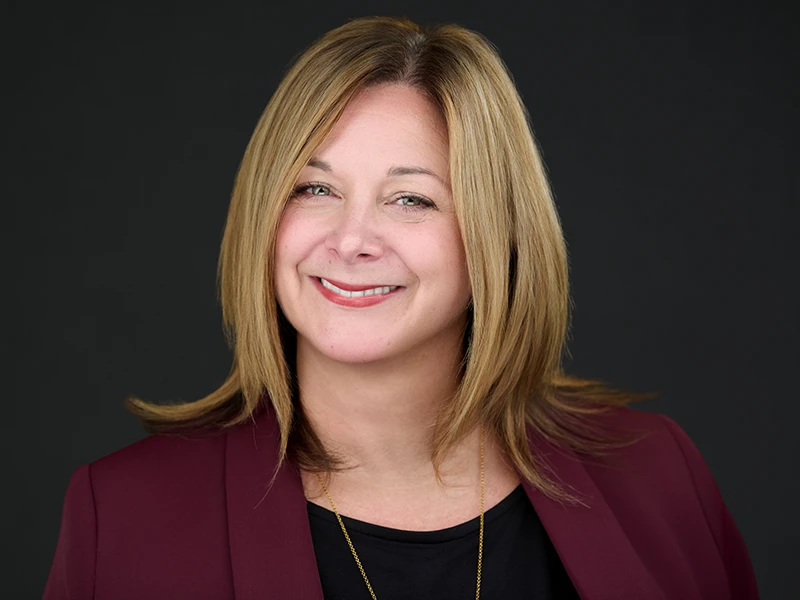Smiling blonde woman in a burgundy blazer, photographed on a dark grey background.