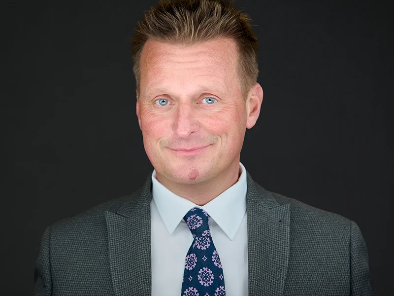 Man in a grey suit with a patterned tie, photographed on a dark grey background.