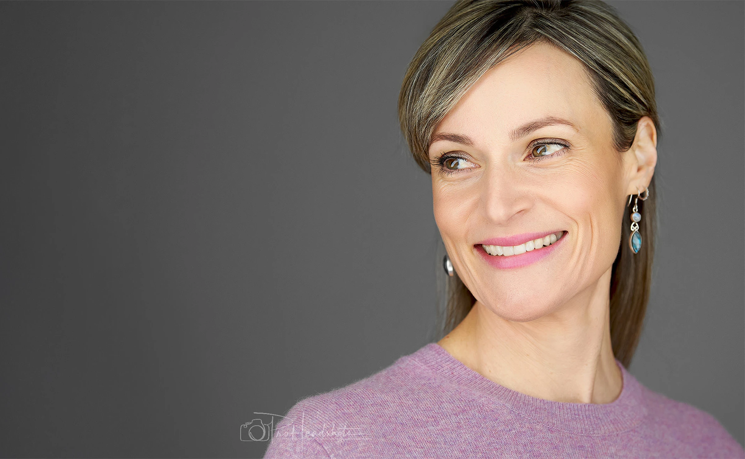 Professional headshot of a woman wearing a mauve sweater and blue drop earrings, showing a natural, engaging smile against a dark background. Pro Headshots logo overlaid in light grey.