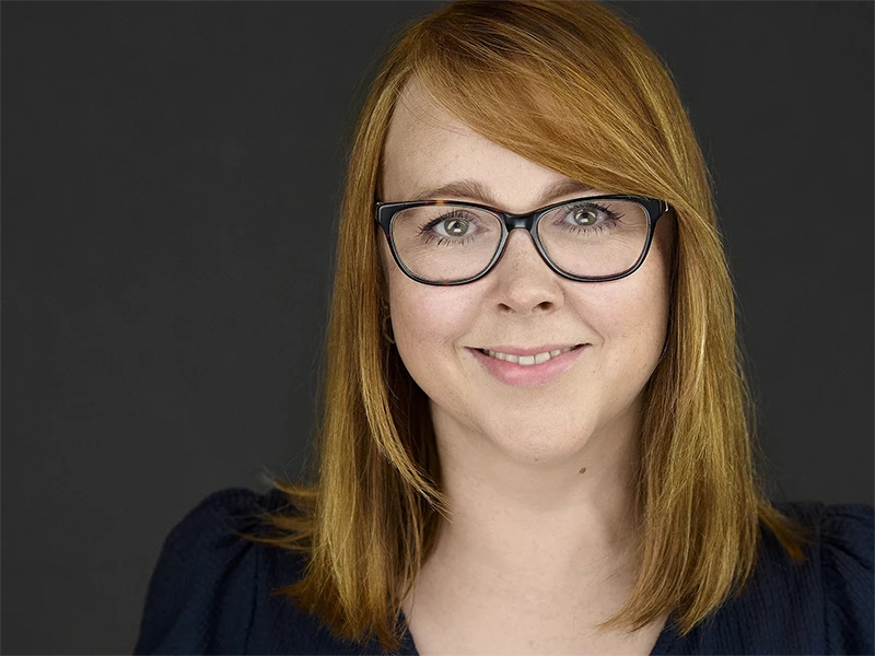 Professional headshot of a woman with red hair wearing glasses and a navy top, showing a friendly, genuine smile against a dark background.