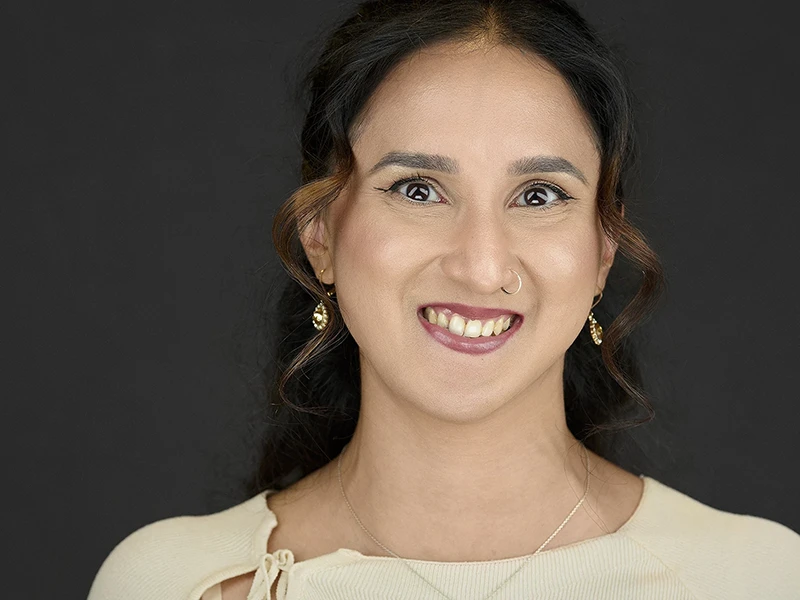 Professional headshot of a woman wearing a cream top with tie detail, gold earrings and nose ring, showing a bright engaging smile against a dark background.