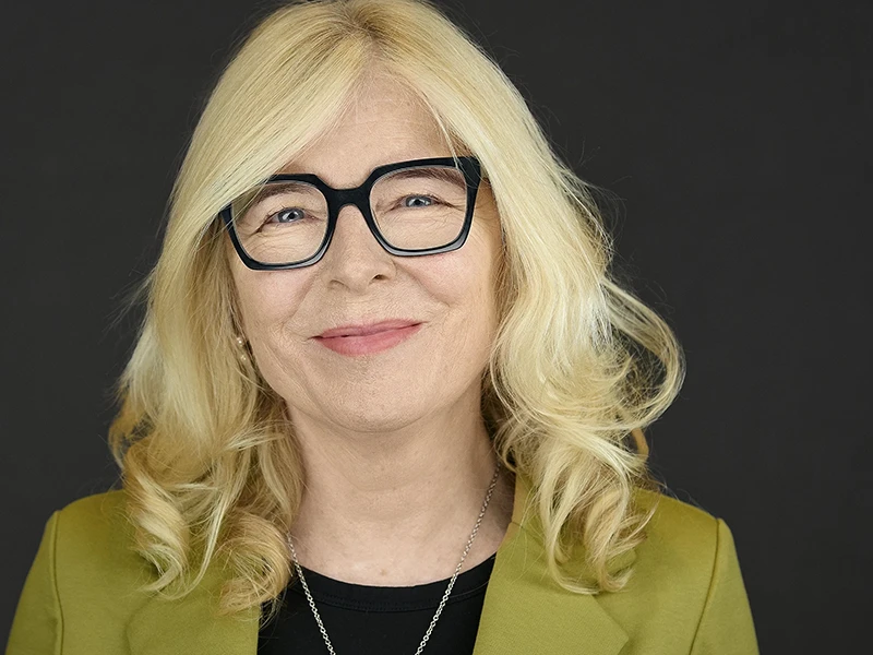 Professional headshot of a woman with blonde curled hair wearing black-framed glasses, a black top and chartreuse blazer, showing a warm confident smile against a dark background.