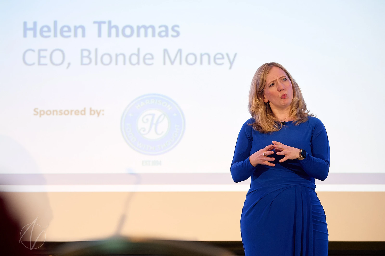 Helen Thomas speaking at a financial conference, her name and title visible on the slide behind her