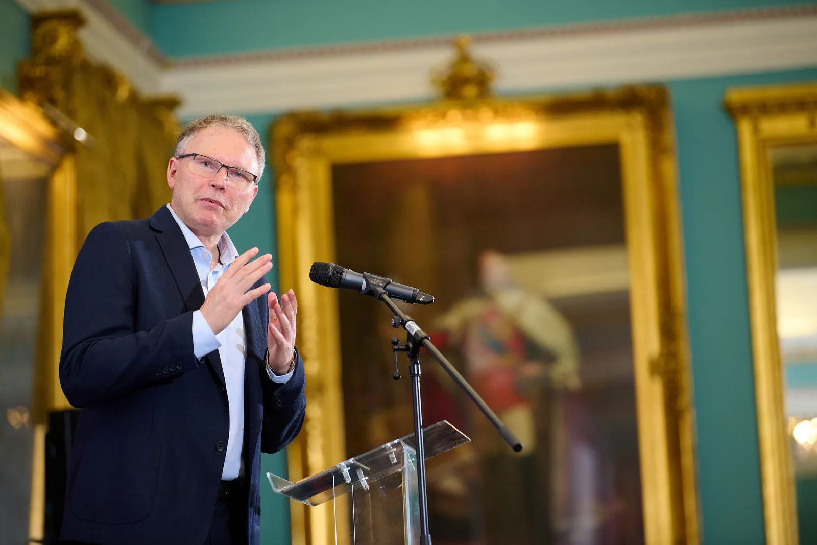 Male speaker at a podium pointing directly at the audience mid-talk, fully engaged at a professional industry event