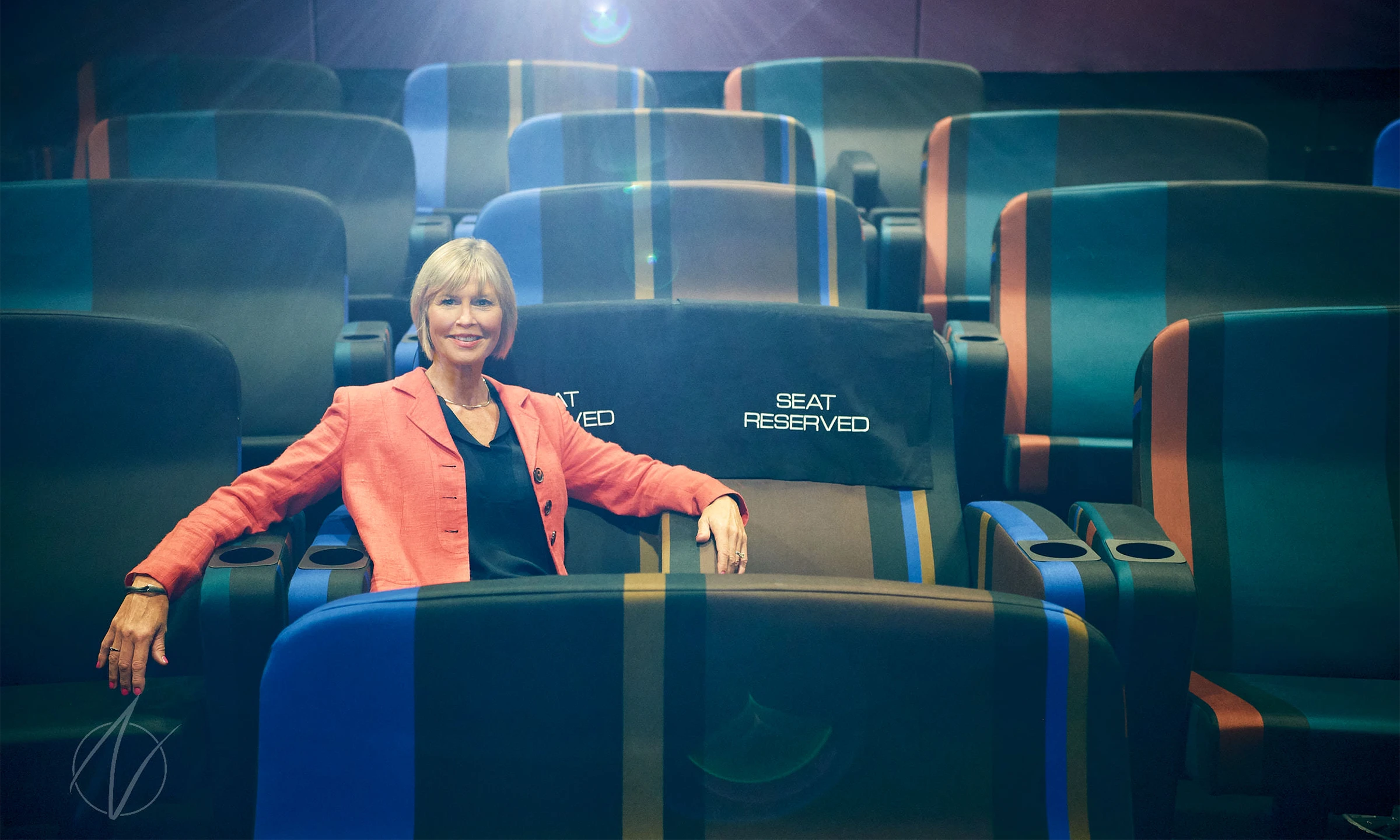 A female speaker in a coral blazer and black top sits confidently among theatre-style reserved seating, showcasing executive presence in a contemporary auditorium with dramatic lighting. The professional portrait captures her approachable expertise while highlighting the speaking venue's modern blue and teal seats.