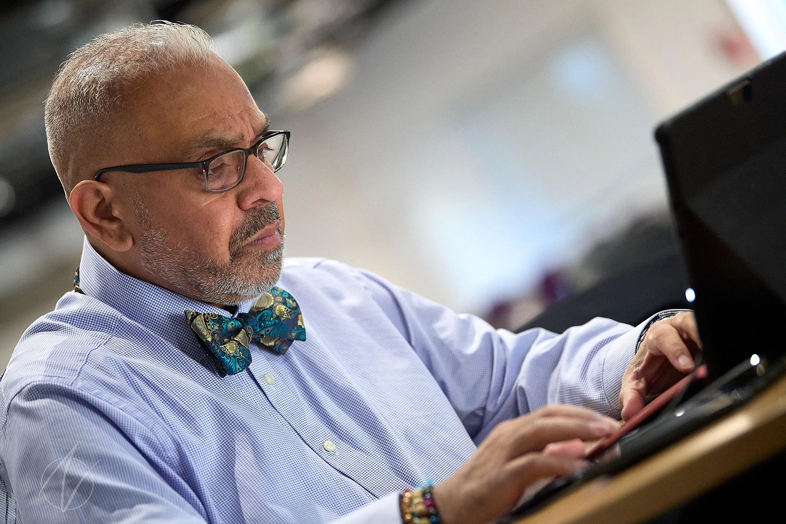 A distinguished professional in a light blue shirt and patterned bow tie concentrates while working at his desk. His thoughtful expression and stylish accessories, including black-framed glasses, create a portrait of focused expertise in action.