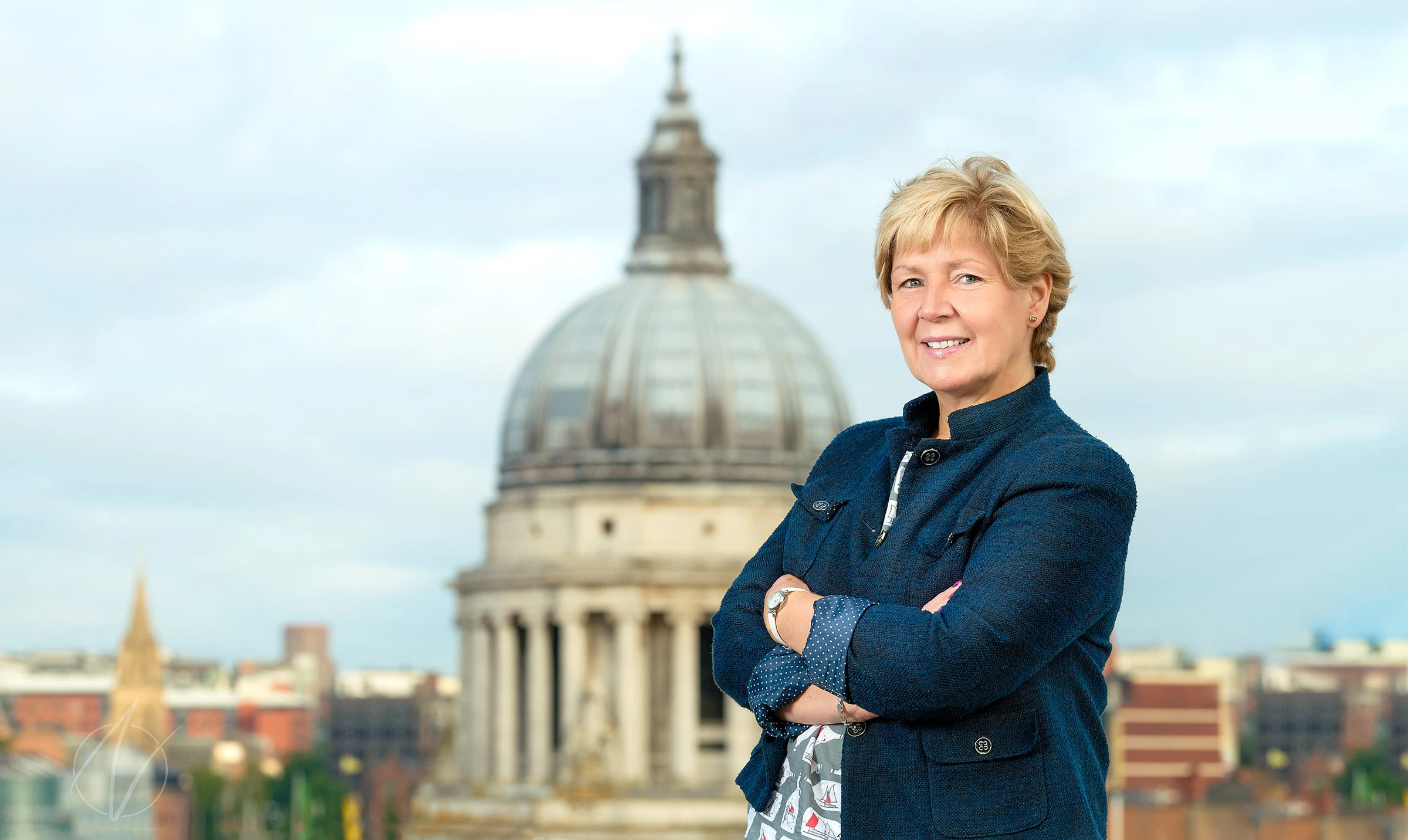 A confident business leader stands with arms crossed in a navy jacket, photographed against the impressive backdrop of a domed cathedral and city skyline. The architectural setting adds gravitas to this outdoor personal branding portrait.