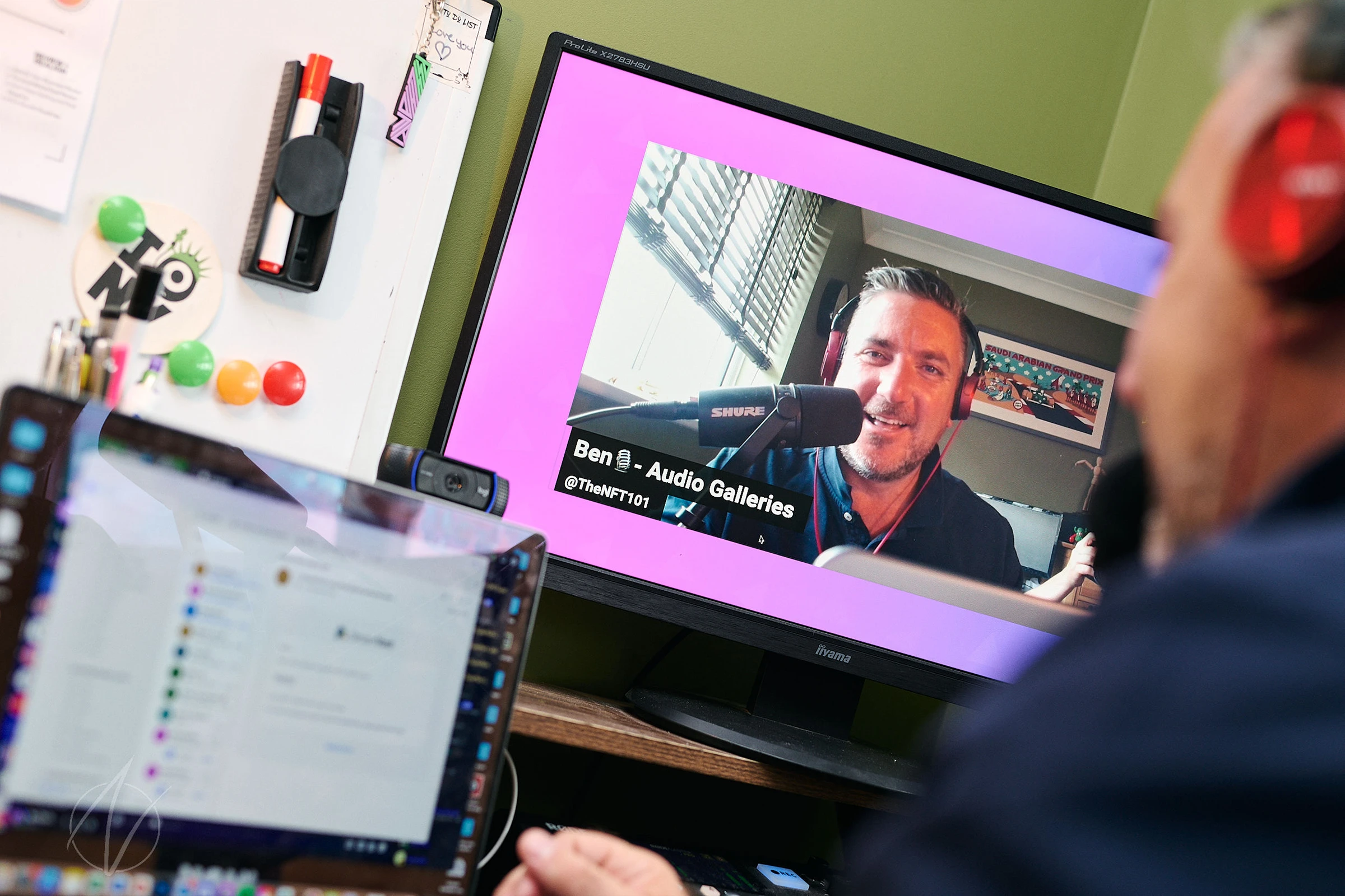 Remote podcast recording setup showing a video call interface on screen with the guest speaker visible, while production notes and colourful markers are arranged on the desk. The image captures the modern digital workspace of a content creator.