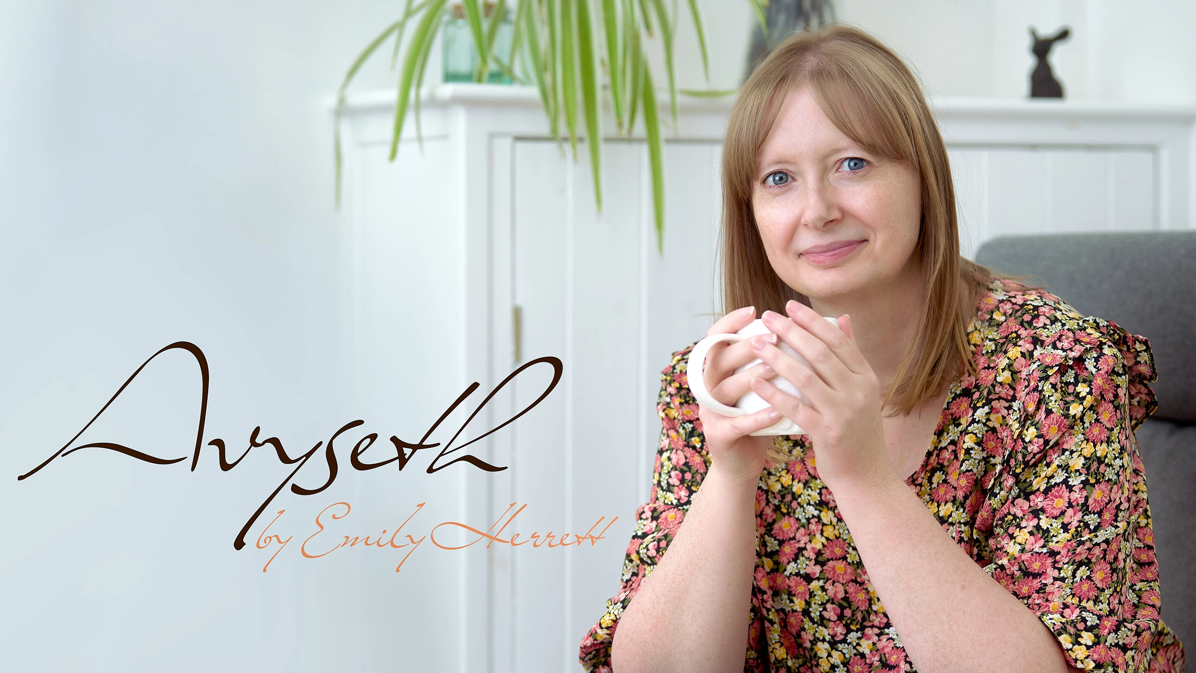 A woman sits comfortably in a grey armchair, holding a white mug with both hands. She's wearing a floral print dress and has straight shoulder-length hair with bangs. Behind her is a white panelled wall with a hanging plant and small decorative statue. Below the image is a stylised signature reading 'Anyseth by Emily Sherratt' in black and coral colours.