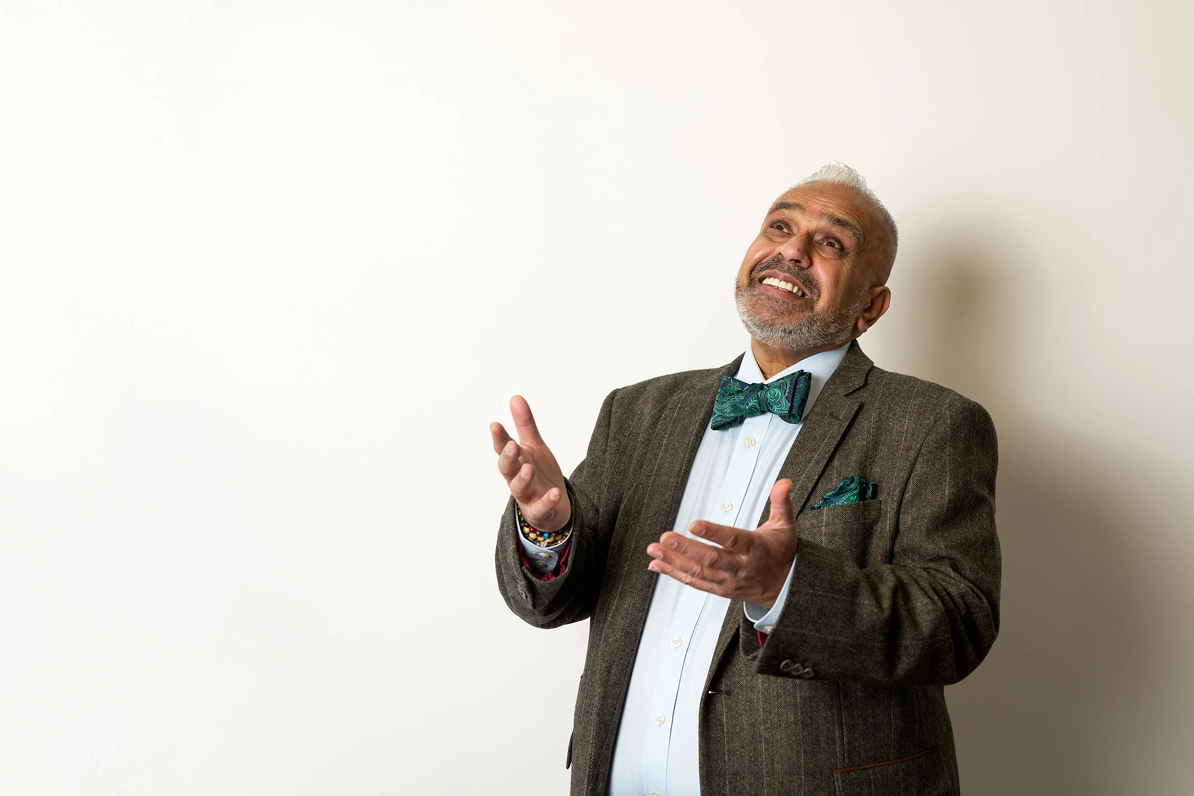 A man in a grey pinstriped suit and teal bow tie gestures animatedly with both hands raised, captured mid-conversation against a white background. His genuine smile and dynamic pose convey warmth and enthusiasm. His outfit is completed with a matching teal pocket square, white dress shirt, and his silver hair and beard add distinguished character to his professional appearance.