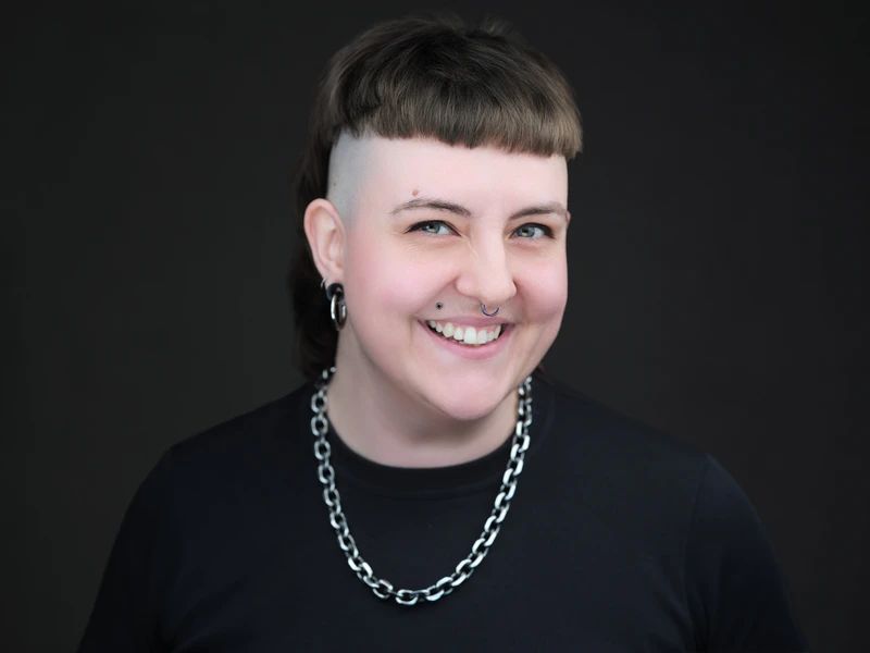 Corporate headshot of a person with short hair wearing a chain necklace against a dark background, smiling.
