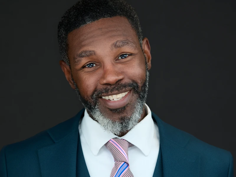 Formal headshot of a man in a navy suit and tie on a dark background
