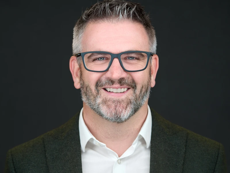Studio-style headshot of a man with glasses on a dark background