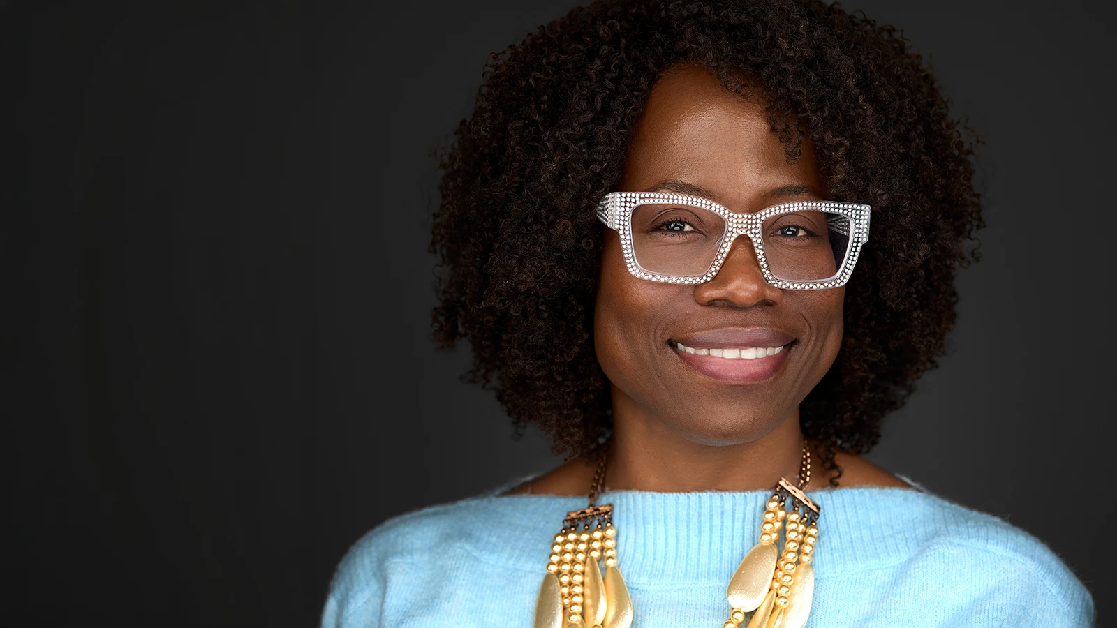 Professional woman with natural curly hair and rhinestone glasses smiling warmly against a dark background, photographed by a Bristol headshot photographer.