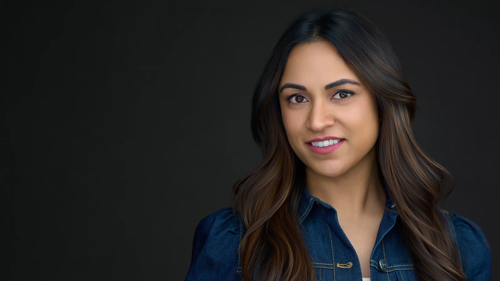 Woman with long dark wavy hair and warm smile in a navy denim jacket, professional headshot by a Devon headshot photographer