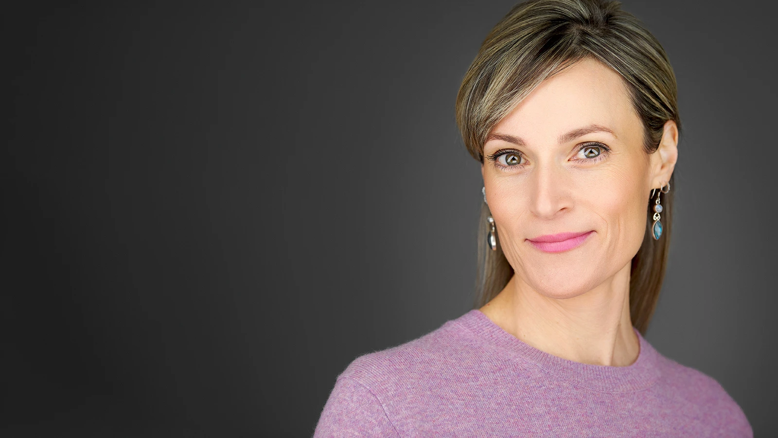 Woman with silver-streaked hair and a composed smile in a lavender sweater, professional headshot on a dark grey background, taken during a Dorset headshot photography session