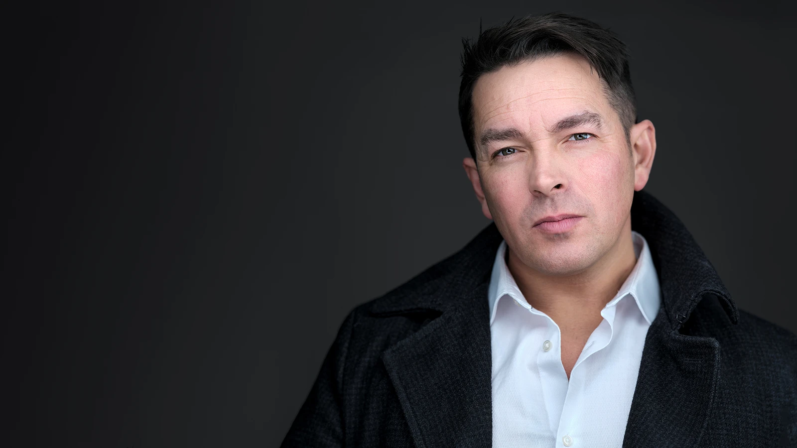 Man with dark hair in dark overcoat and white open-collar shirt, calm confident expression, professional headshot by Falmouth photographer