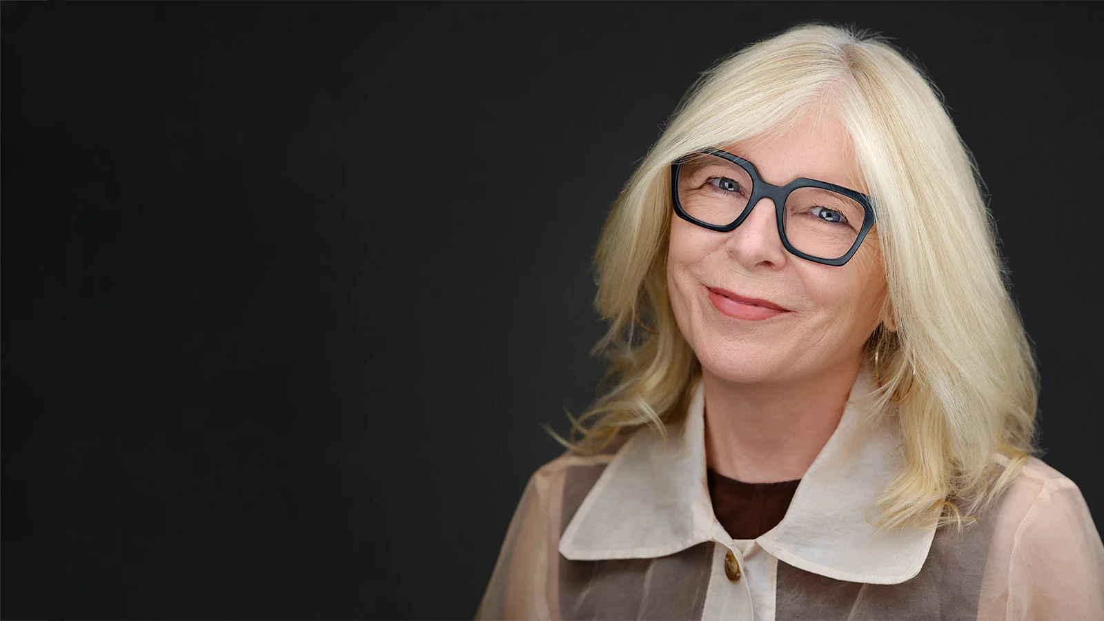 Business headshot of a smiling woman with blonde hair and dark-rimmed glasses against a charcoal studio background