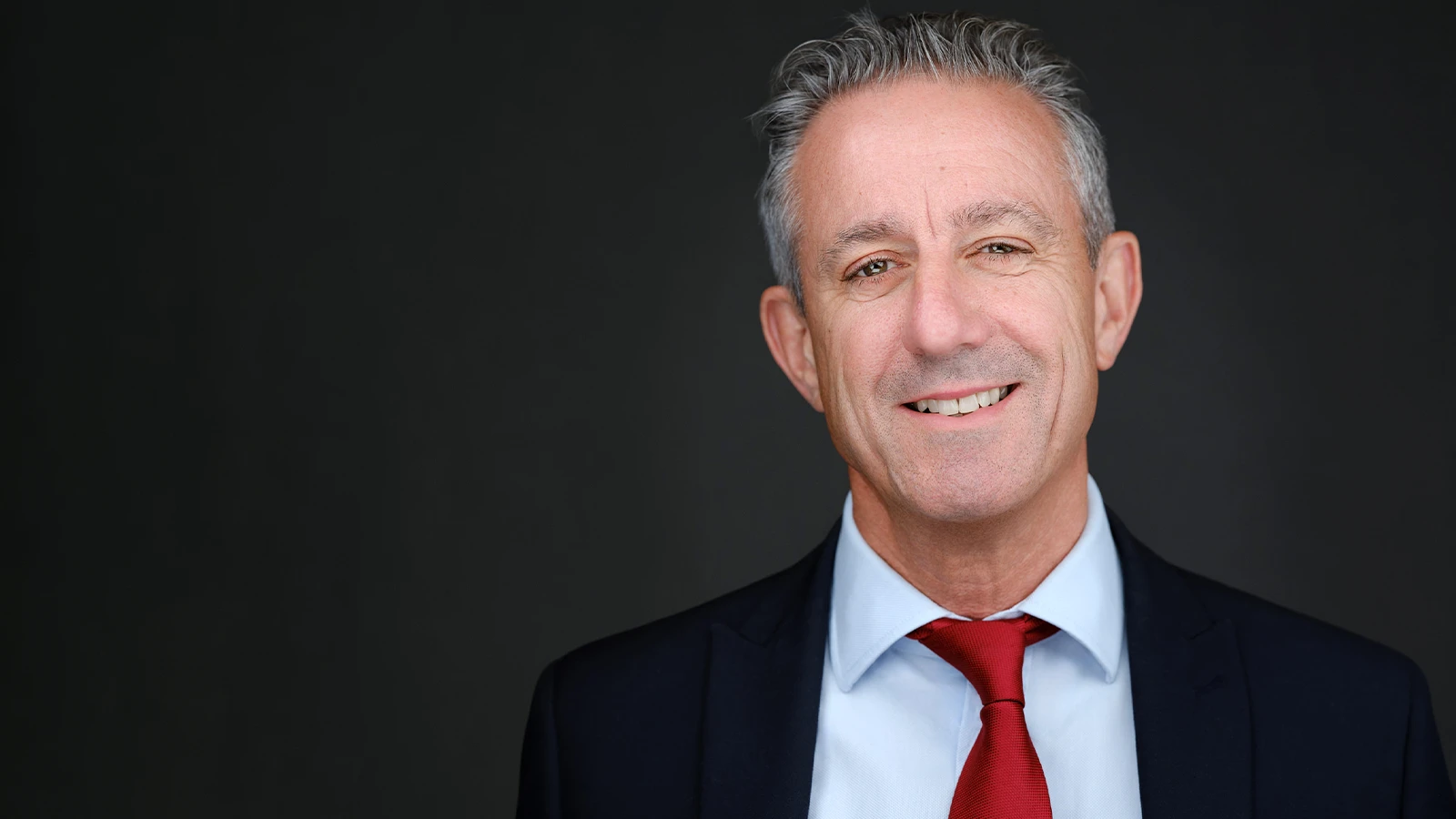 Corporate headshot of a man with grey hair in a navy suit and red tie, warm relaxed smile