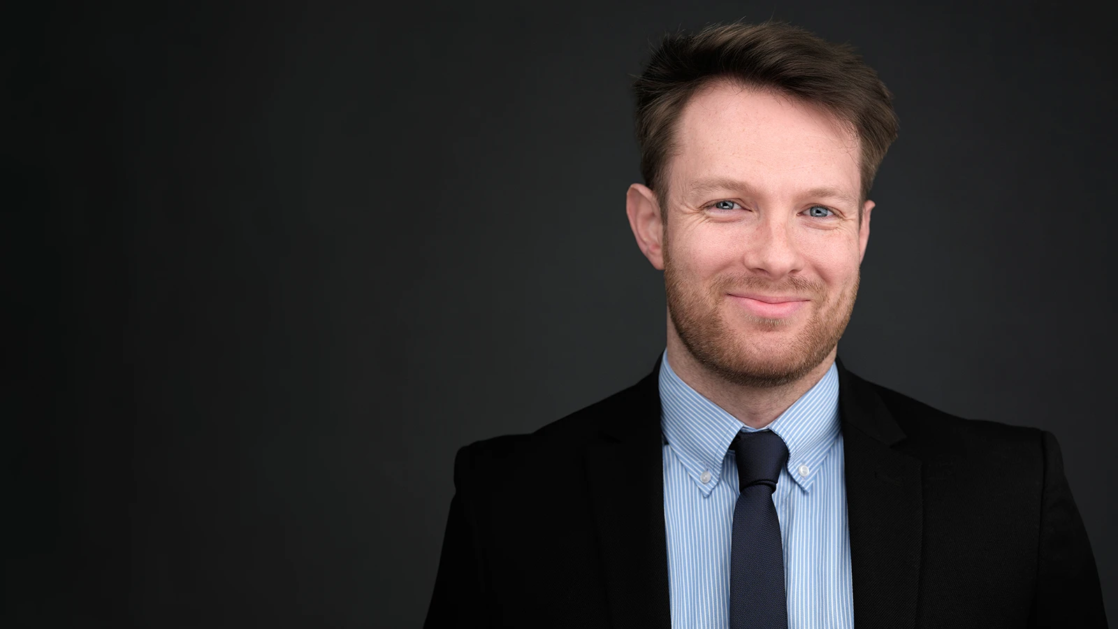 Man in a dark suit and blue striped shirt, professional headshot on a charcoal studio background